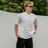 Man wearing a white t-shirt and black pants standing outdoors against a white wall with greenery.